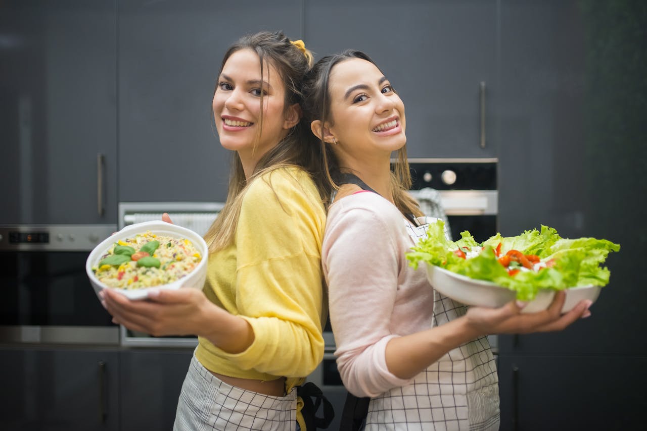 Two smiling women hold vibrant salads in a kitchen setting, showcasing fresh culinary creations.